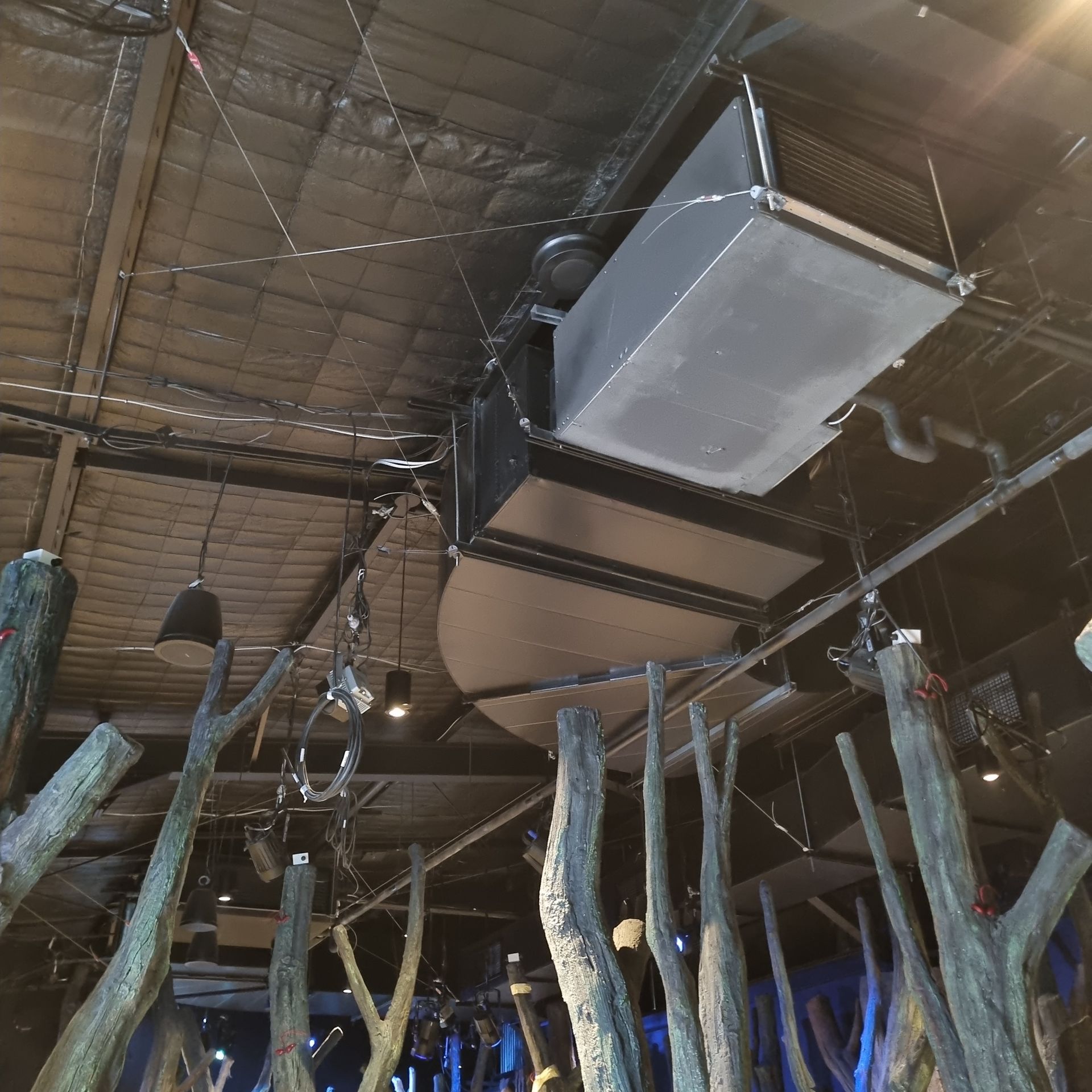 Indoor ceiling with large silver HVAC ducting above rustic wooden posts and hanging lights.