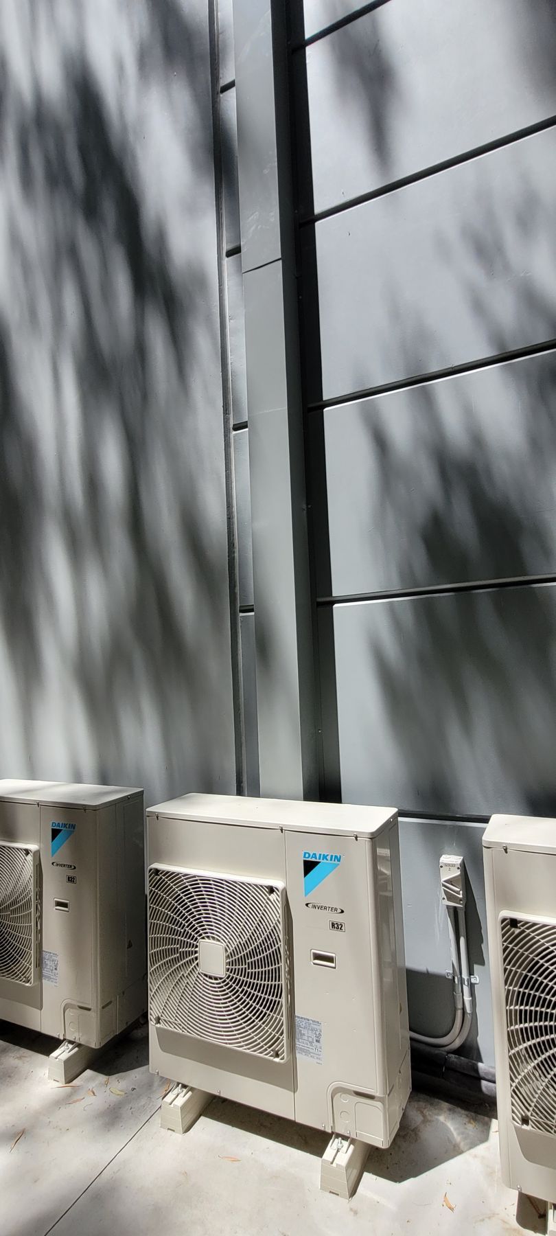 Row of outdoor air-conditioning units beside a gray wall with dappled sunlight and shadows
