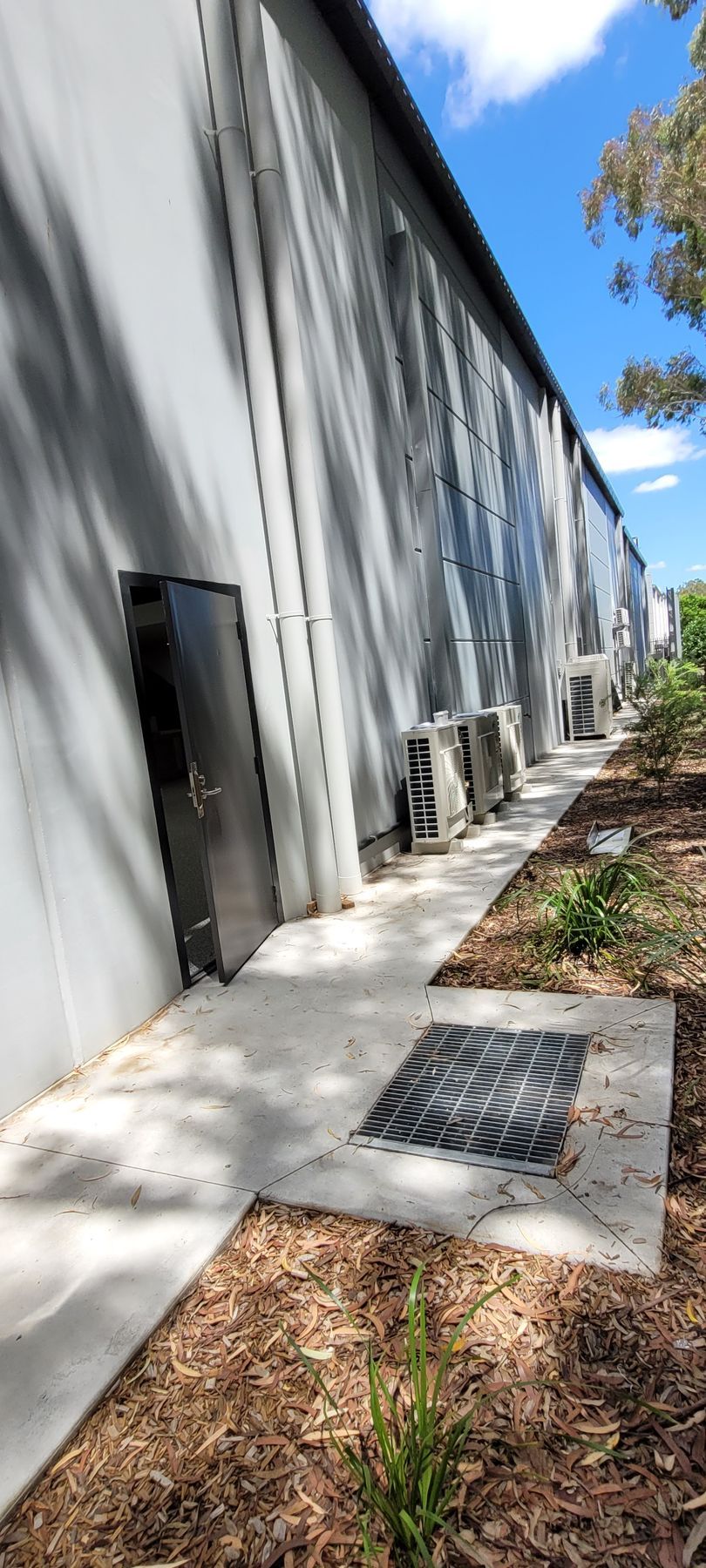 Long white building exterior with windows, concrete walkway, and trees along the side under a blue sky