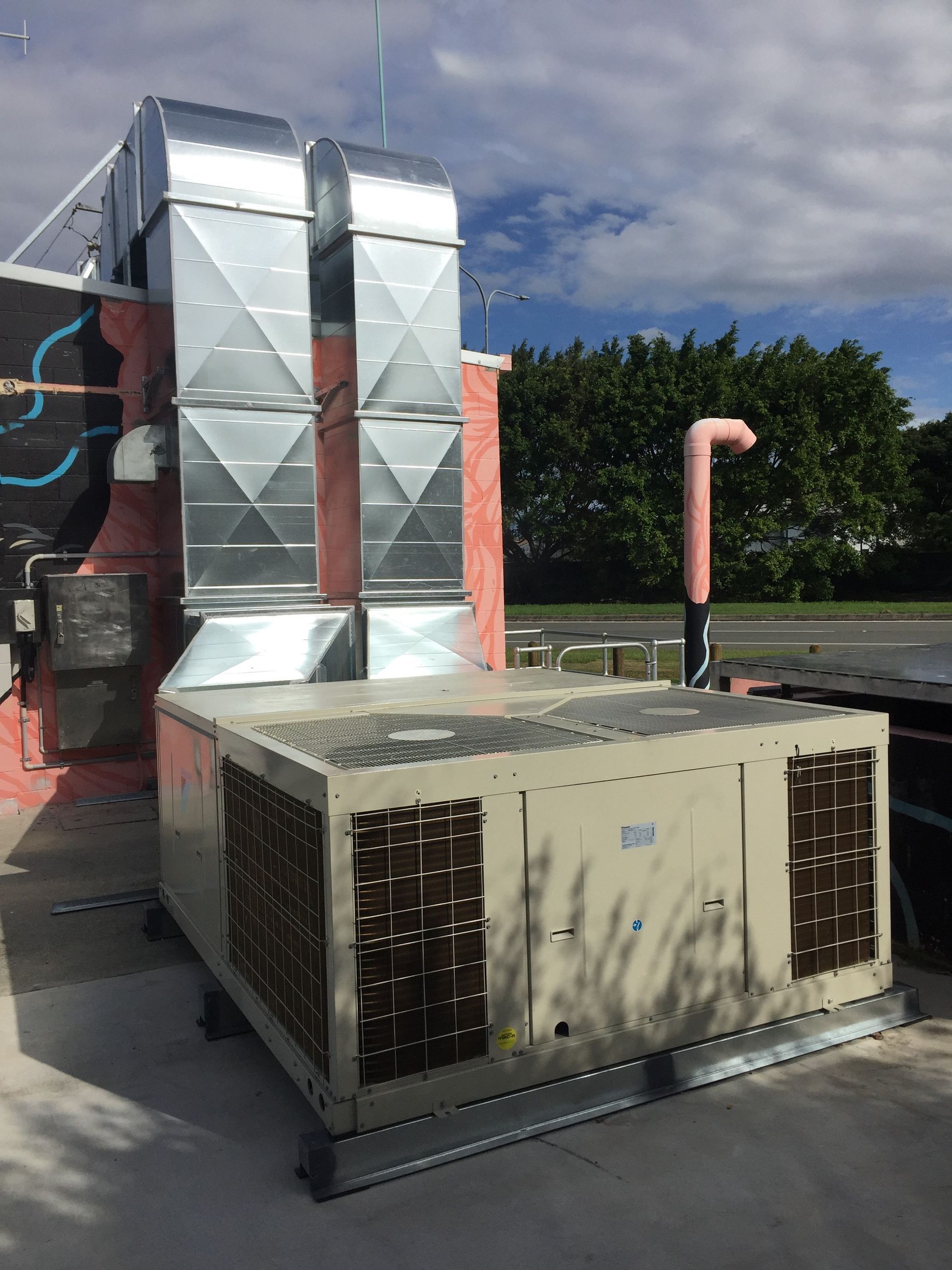 Industrial rooftop HVAC units beside a brick wall, with metal ductwork and trees in the background