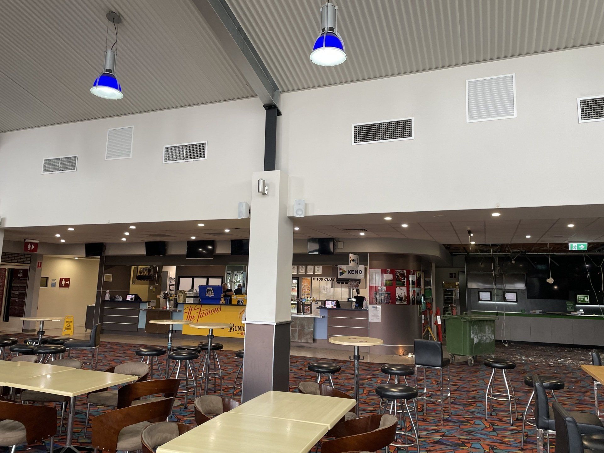 Empty cafeteria with tables and chairs under blue hanging lights, with a counter in the background
