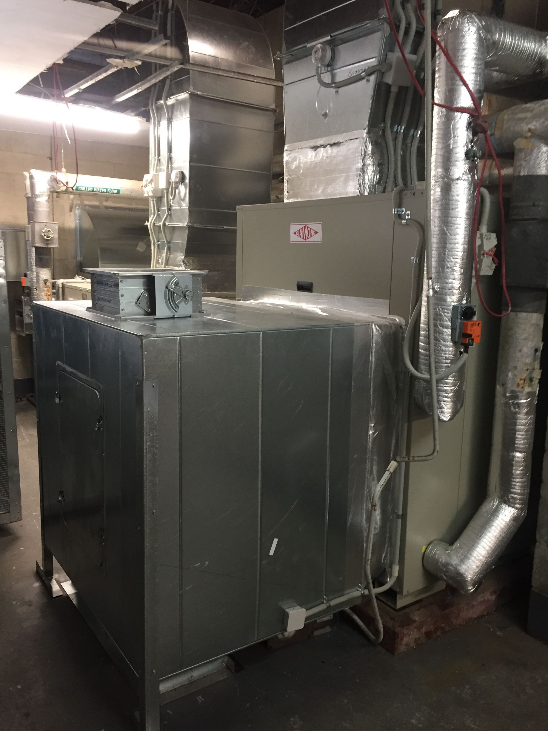 Large metal HVAC units and ductwork in a utility room with exposed ceiling pipes.