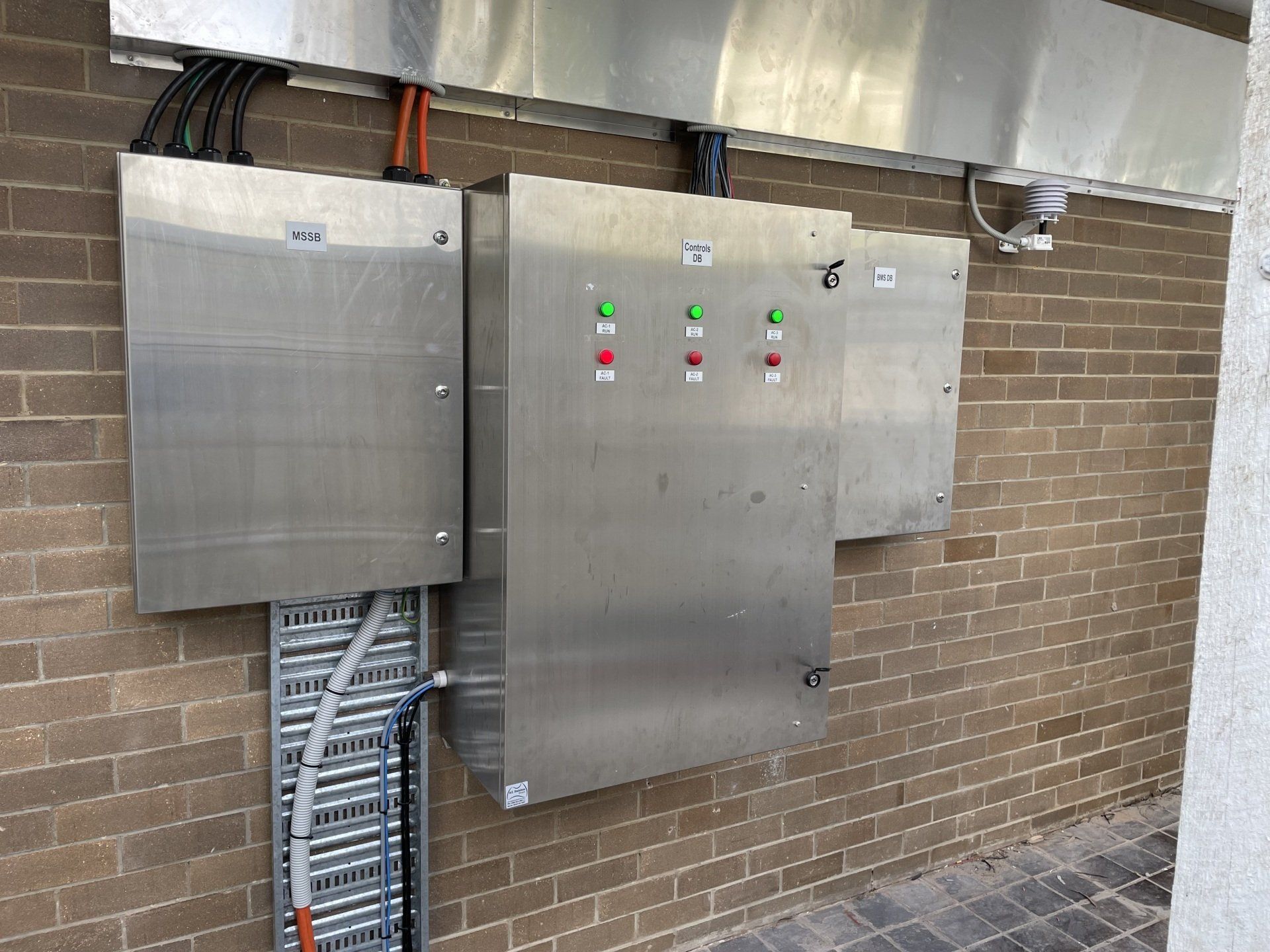 Two stainless steel electrical control panels mounted on a brick wall with colored indicator lights.