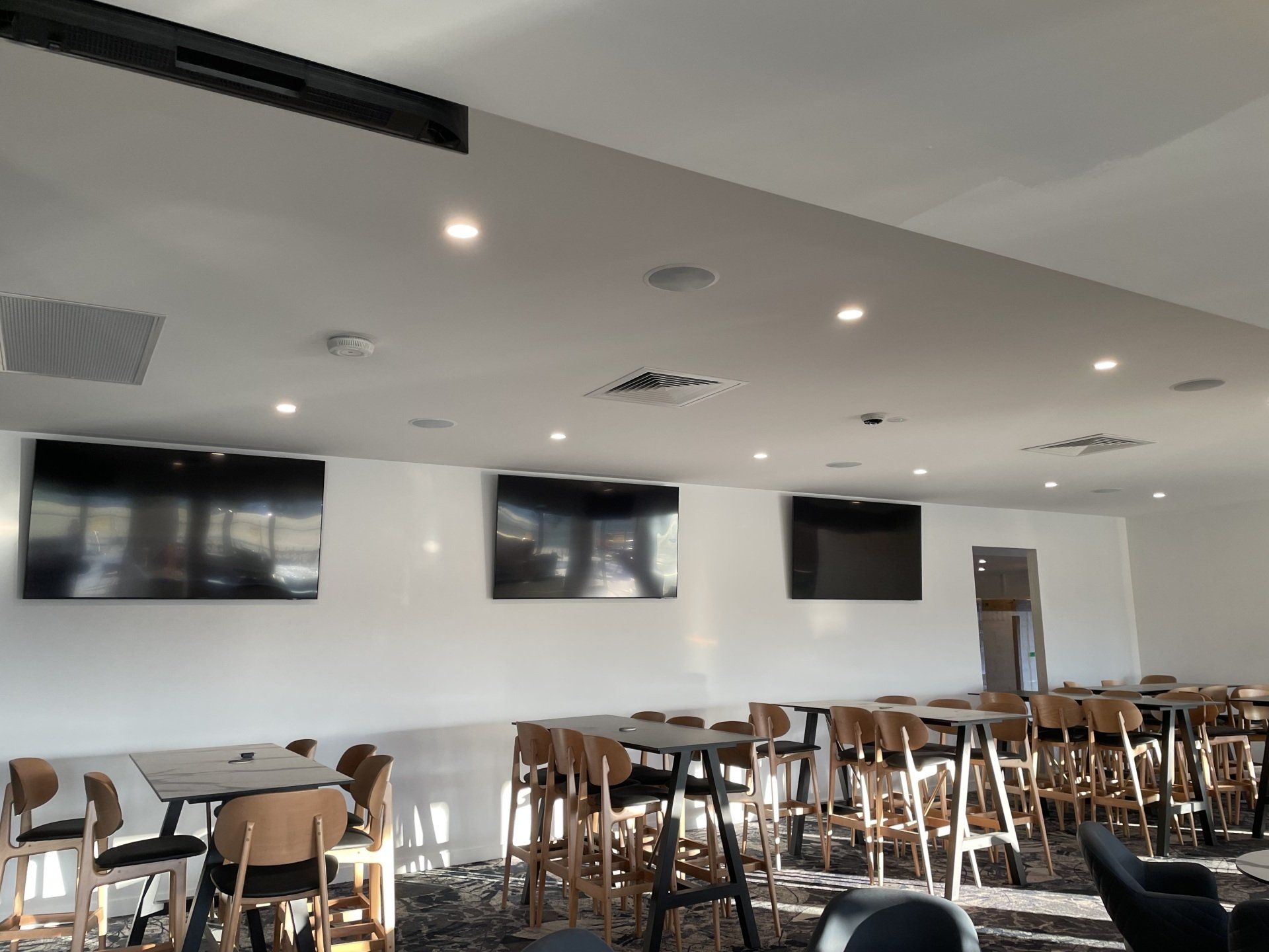 Modern restaurant dining room with wooden chairs, tables, wall-mounted TVs, and recessed ceiling lights