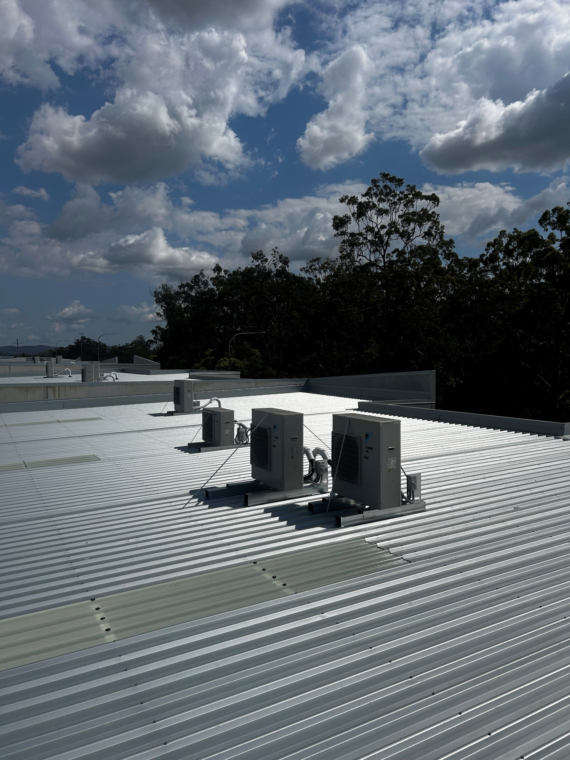 Corrugated metal rooftop with HVAC units under a cloudy blue sky, trees in the background
