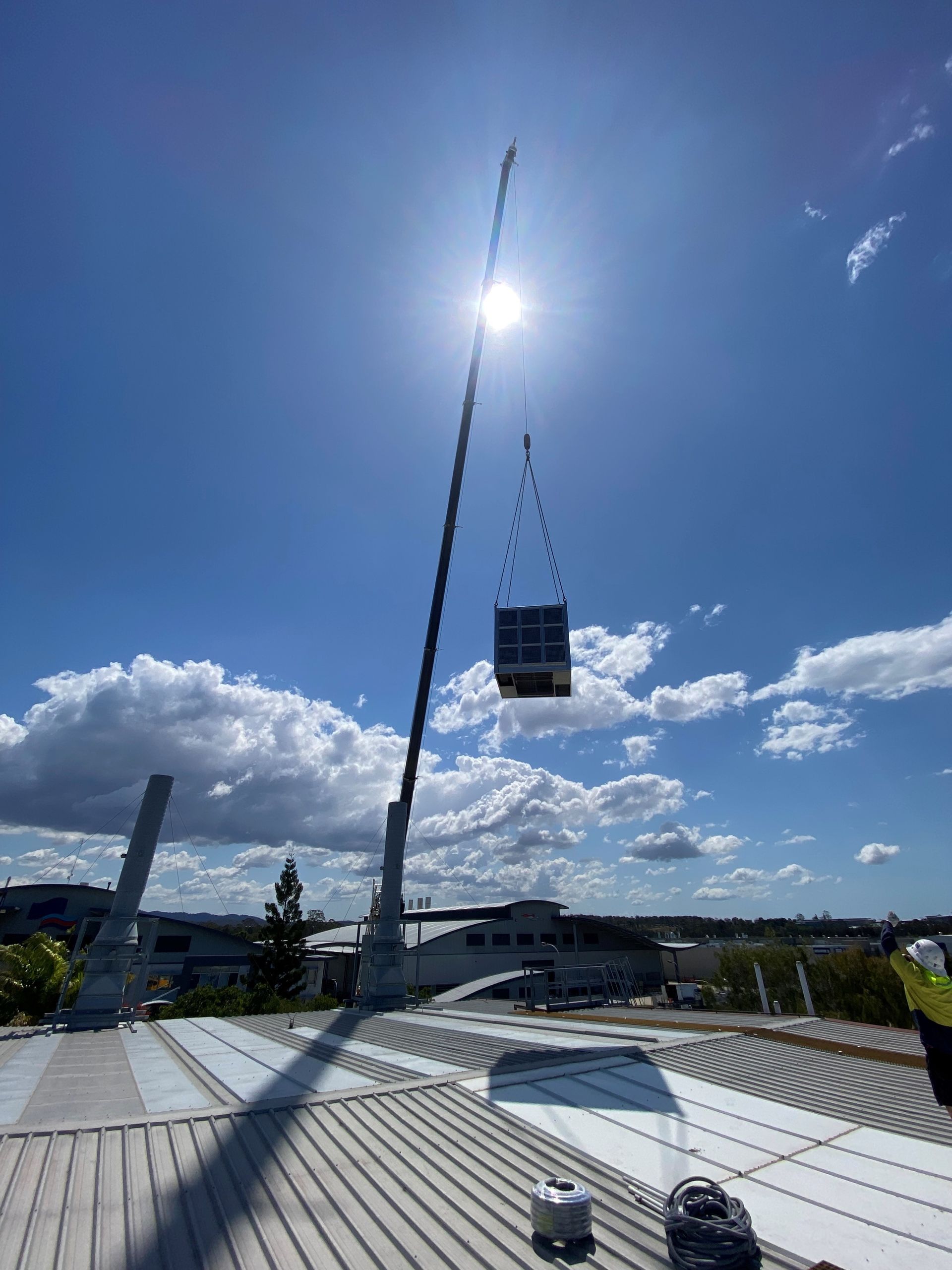 Crane lifting a cube over a white rooftop under a bright blue sky with clouds.
