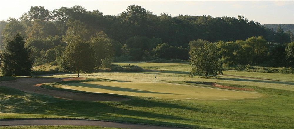 A golf course with green grass, trees, and a sand trap. The sky is clear.