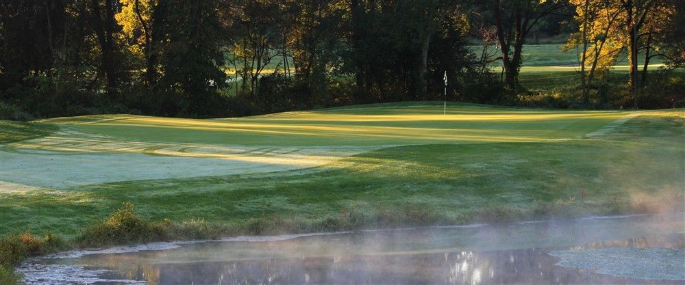 A golf course green with a flag, mist rising from a pond in the foreground, trees in the background.