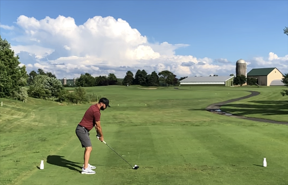 Man in red shirt tees off on a golf course, blue sky with clouds in background.