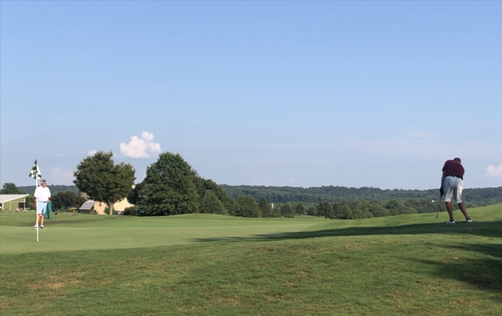 Two golfers on a green golf course on a sunny day; one swings a club, and another stands near the flag.