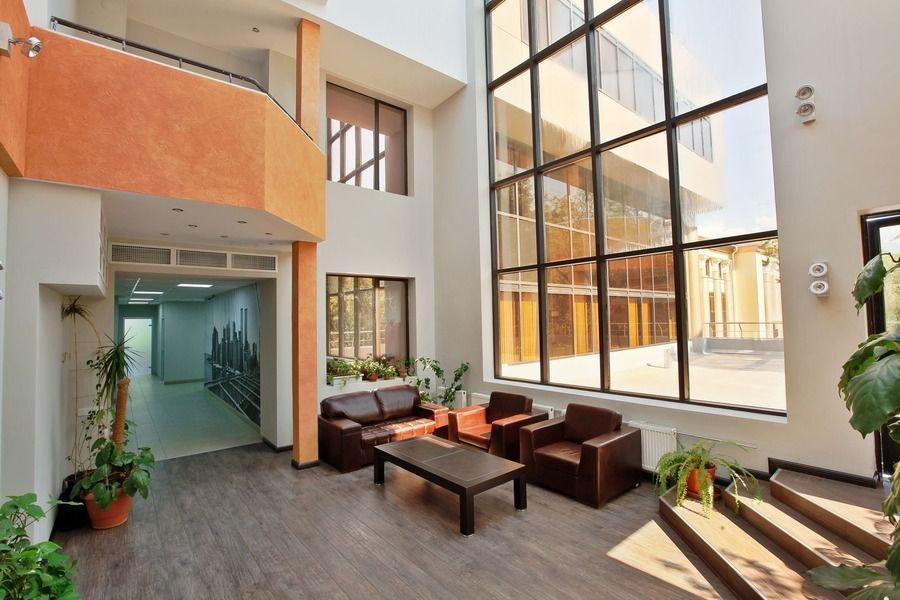 Modern atrium lounge with sofas, coffee tables, plants, and a tall glass wall letting in natural light