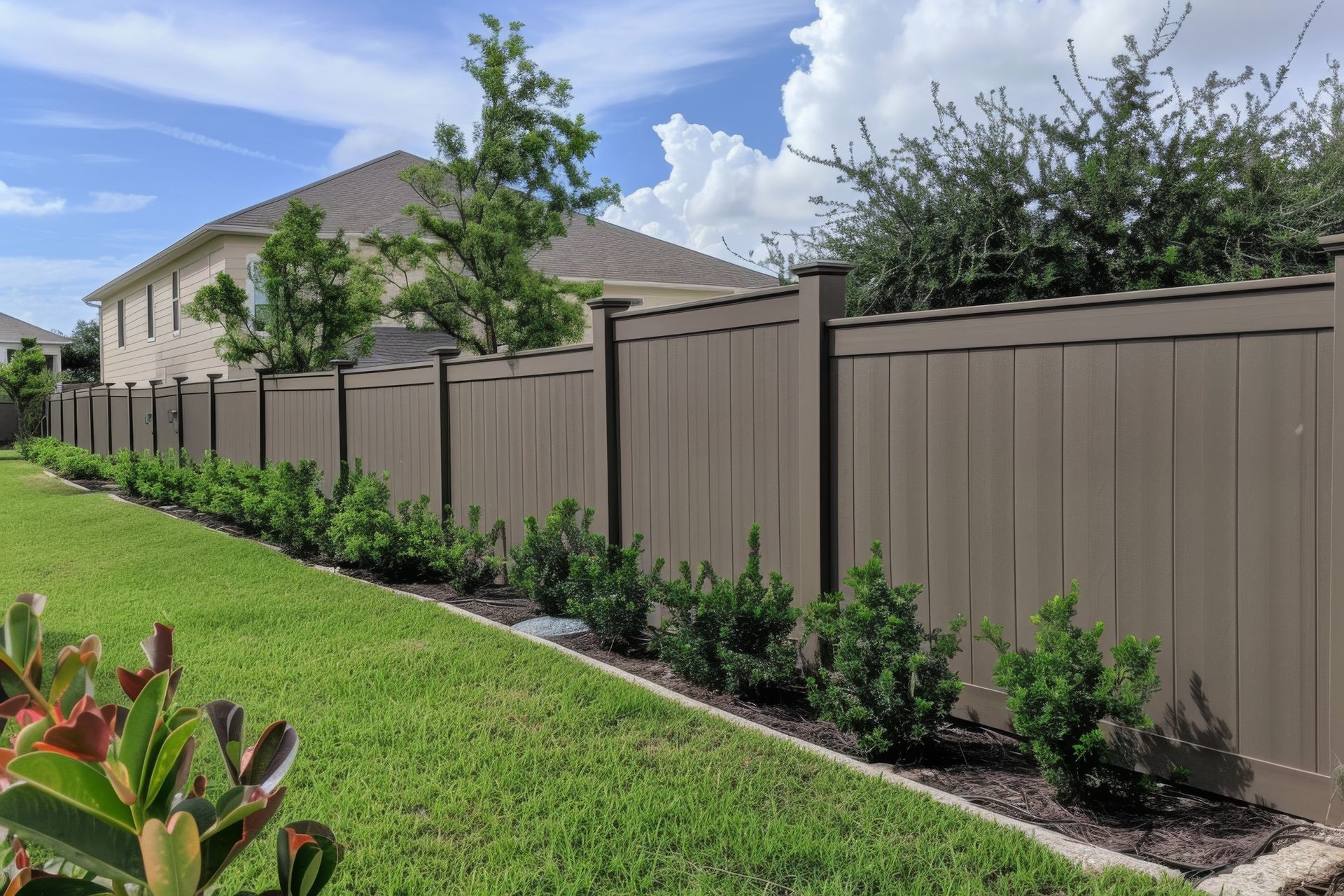 Tan fence lining a green yard with shrubs and a house under a blue sky