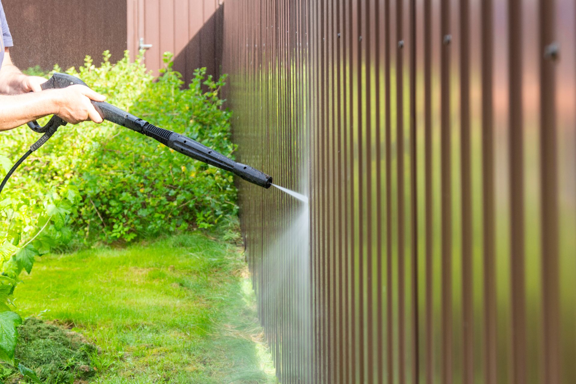 Person pressure washing a brown fence beside a green lawn and shrubs