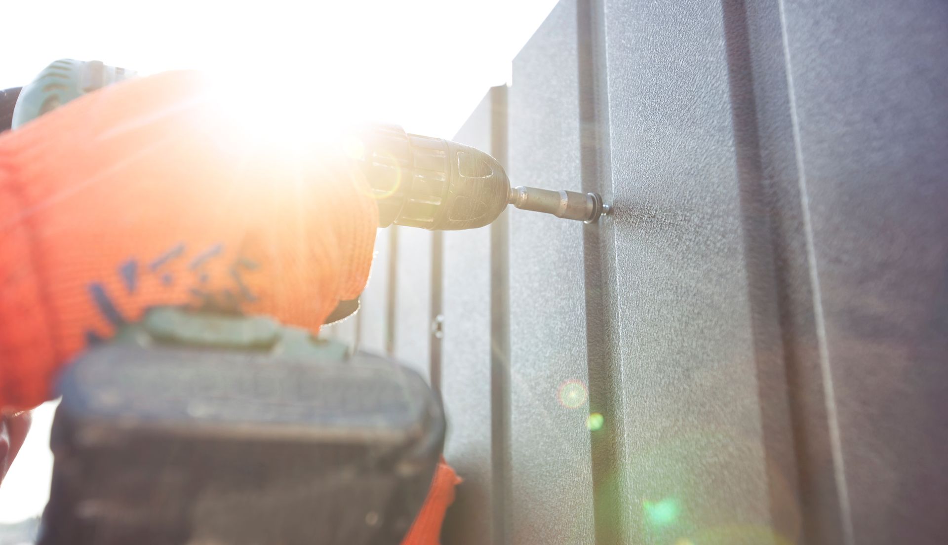 Handheld drill driving a screw into a gray metal fence in bright sunlight