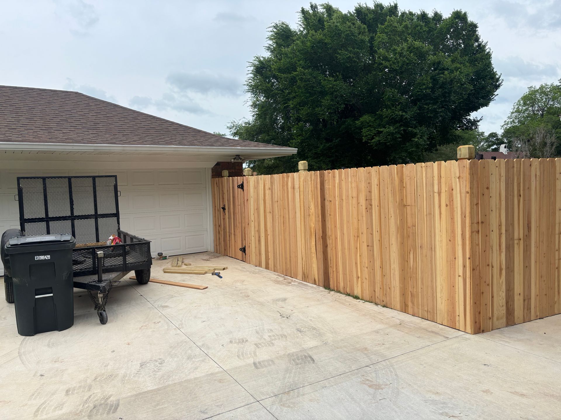 Backyard patio with a new wooden privacy fence beside a house and trash bins on the concrete driveway