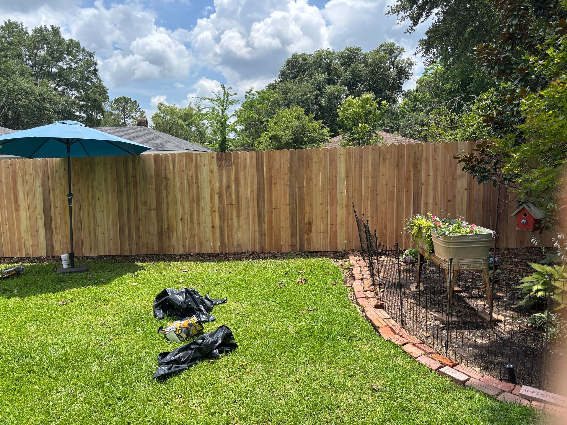 Backyard with green lawn, wooden fence, patio umbrella, and small garden bed under a cloudy sky