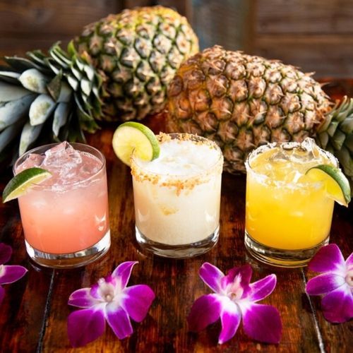 Three drinks are sitting on a wooden table next to pineapples and flowers.