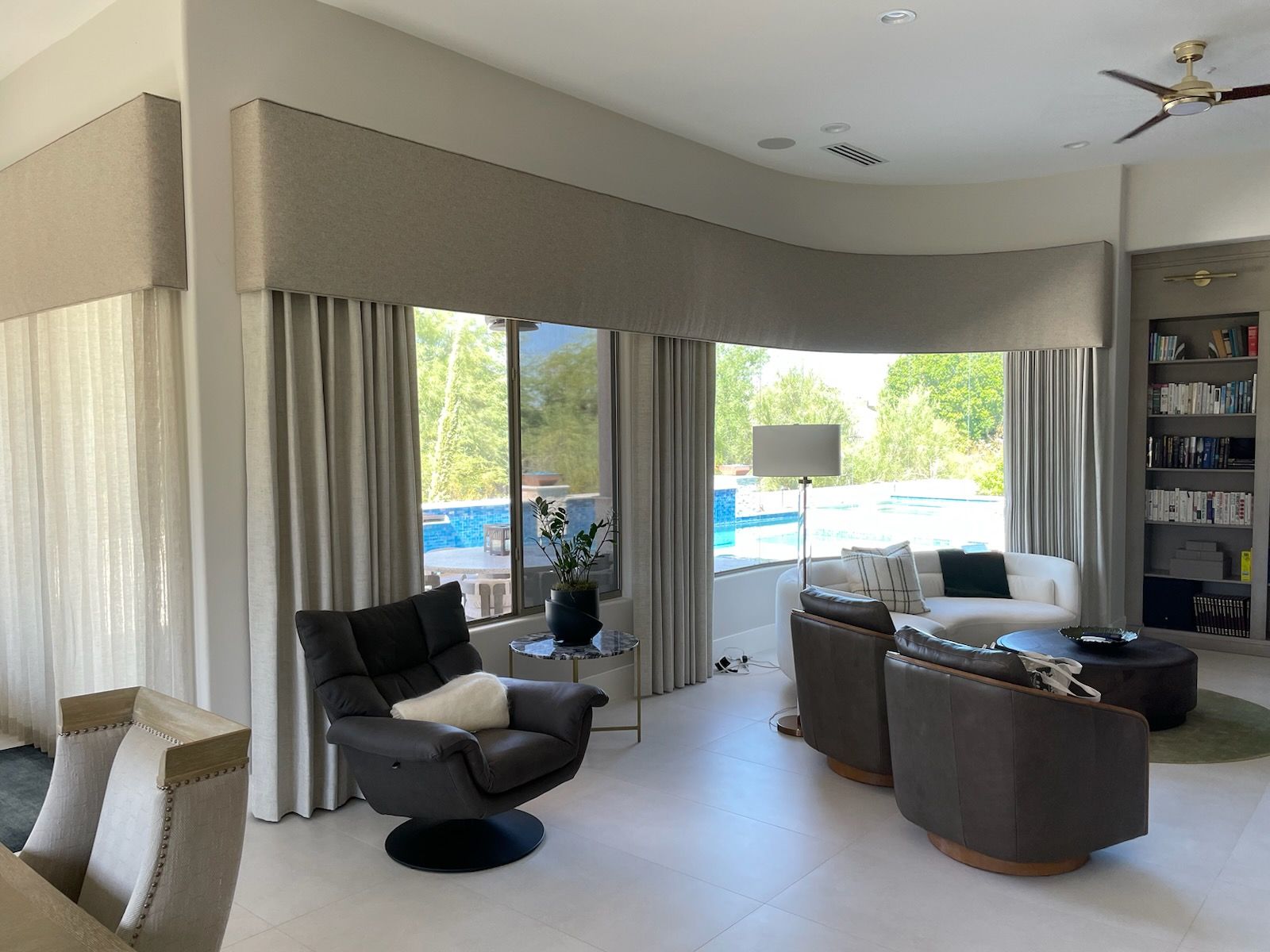 Modern living room with beige floor-to-ceiling curtains, a custom valance, and neutral swivel armchairs facing a pool view.