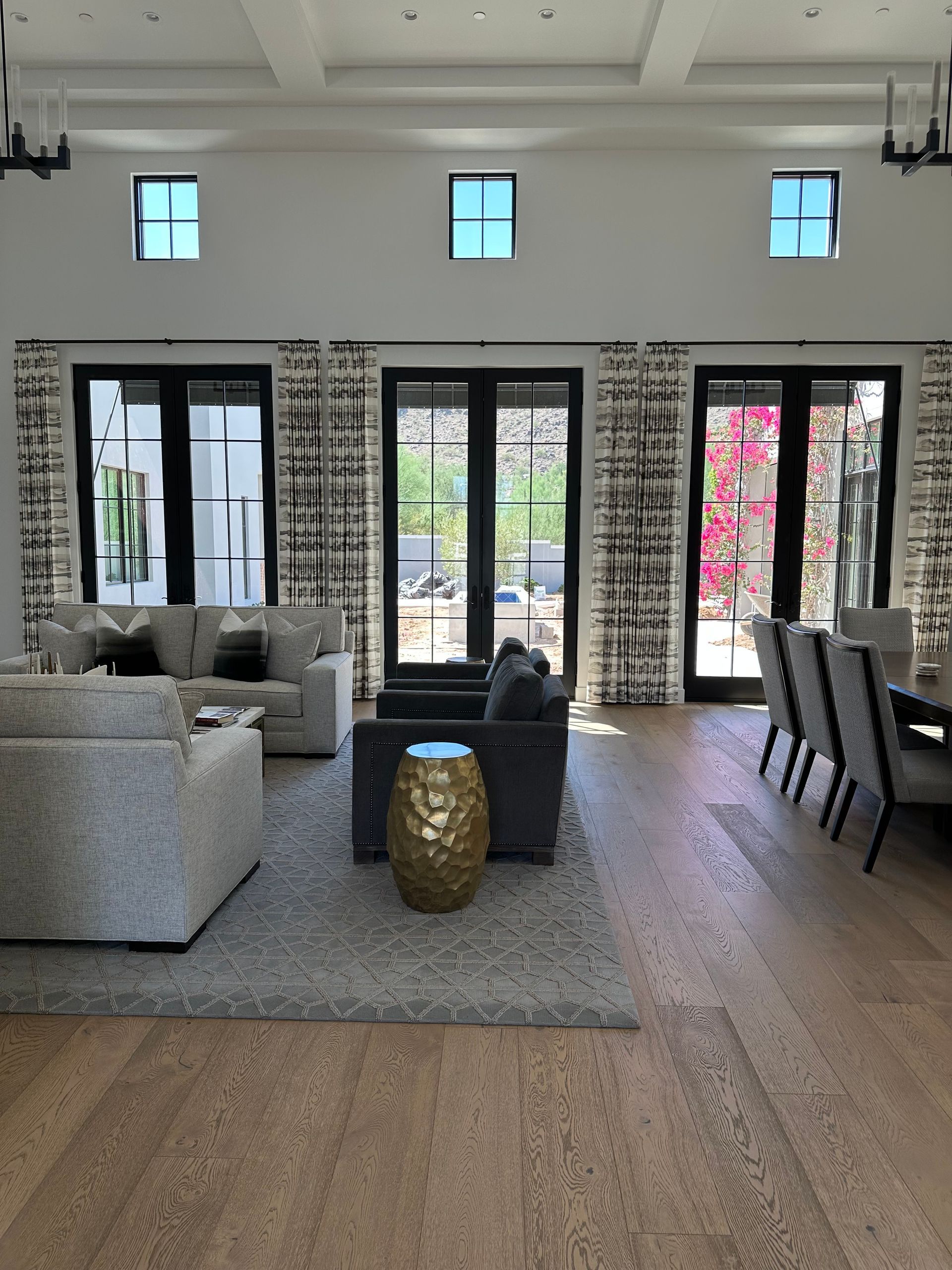 A spacious, modern living room with light wood floors, gray furniture, a gold side table, and large black-framed doors.