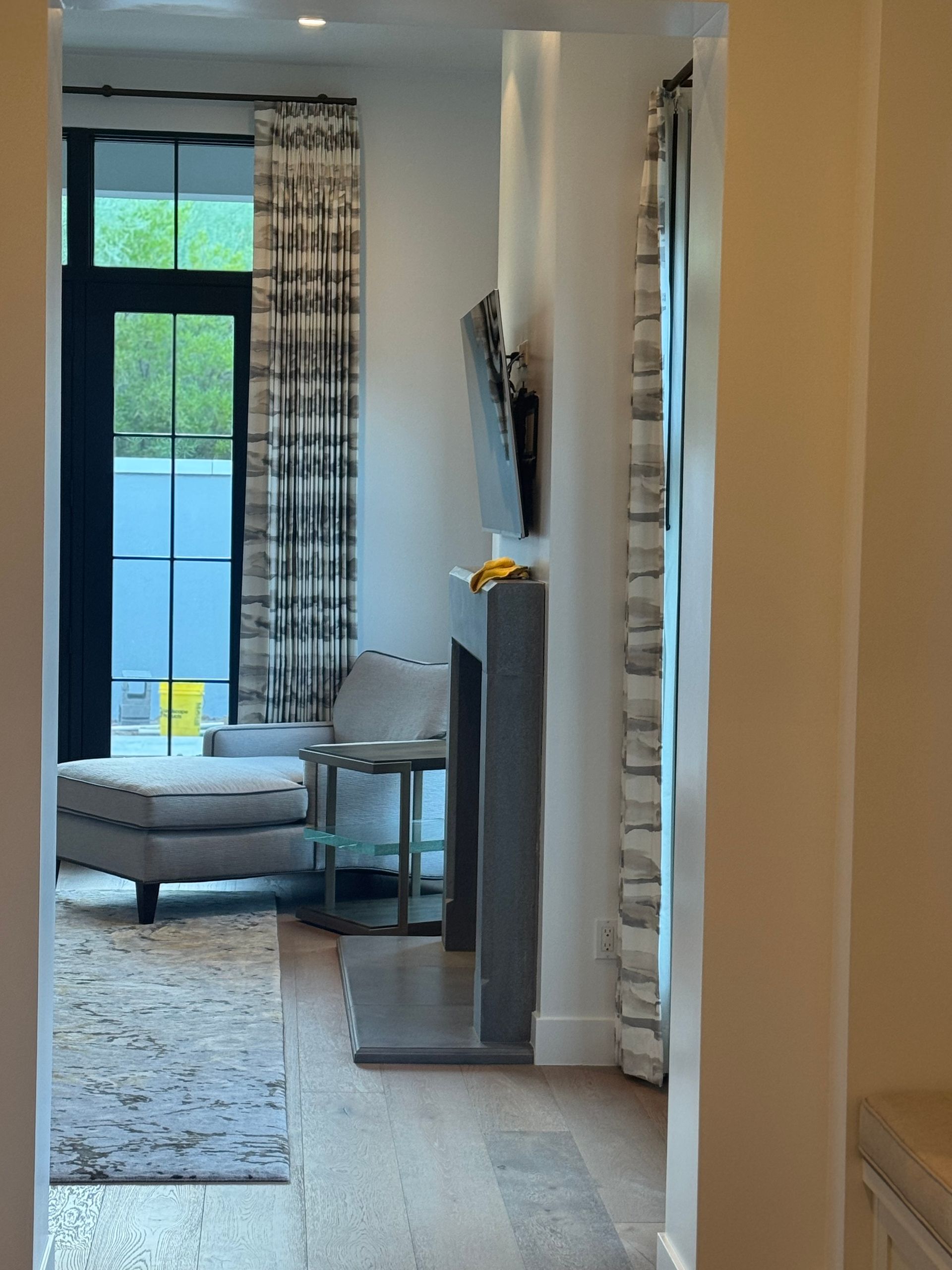 A view through a doorway into a room with a chaise lounge, gray fireplace, wall-mounted television, and floor-length drapes.