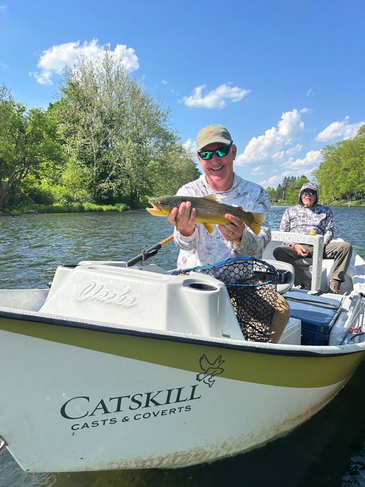 Two anglers in a small boat, one holding up a fish on a sunny river with trees and blue sky.