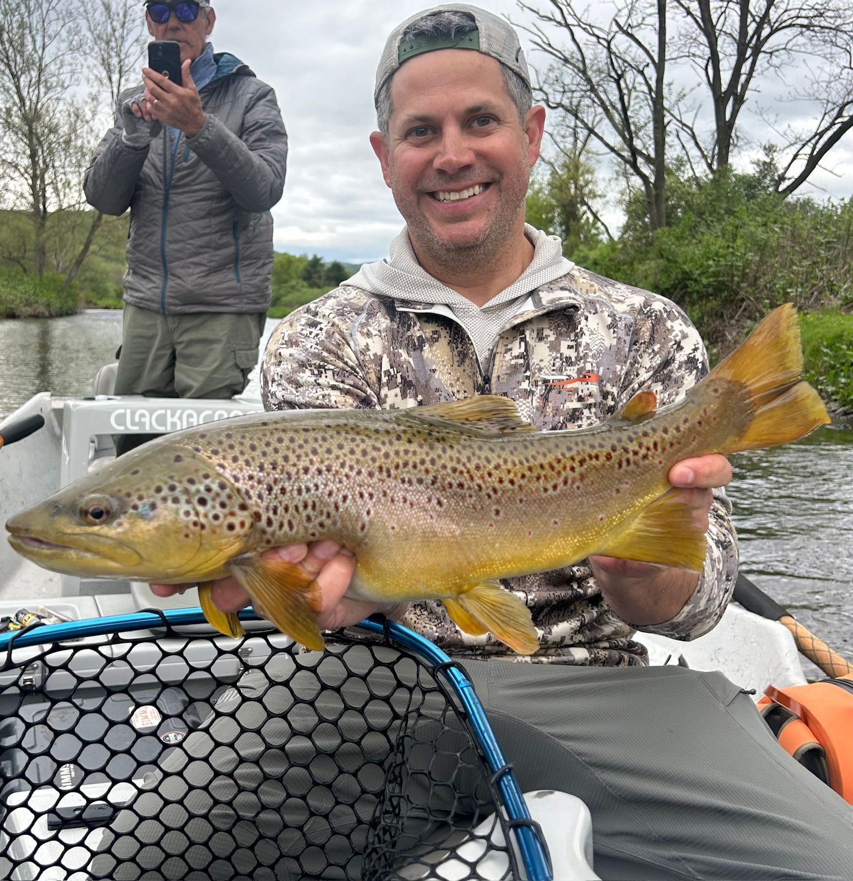 Angler holding a large brown trout on a boat, with another person photographing in the background.