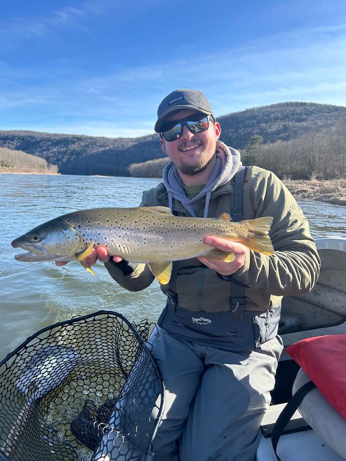 Man in sunglasses holding a large trout on a boat on a lake with mountains in the background