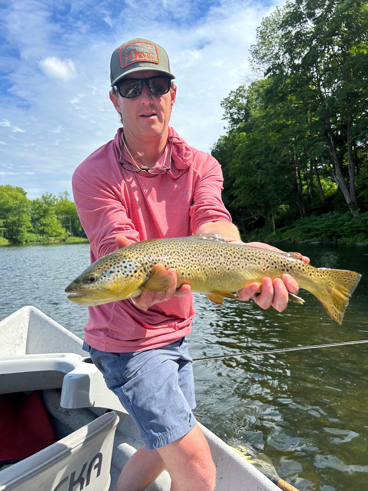 Angler in a boat holding a large trout on a sunny river, wearing a pink shirt and camo cap