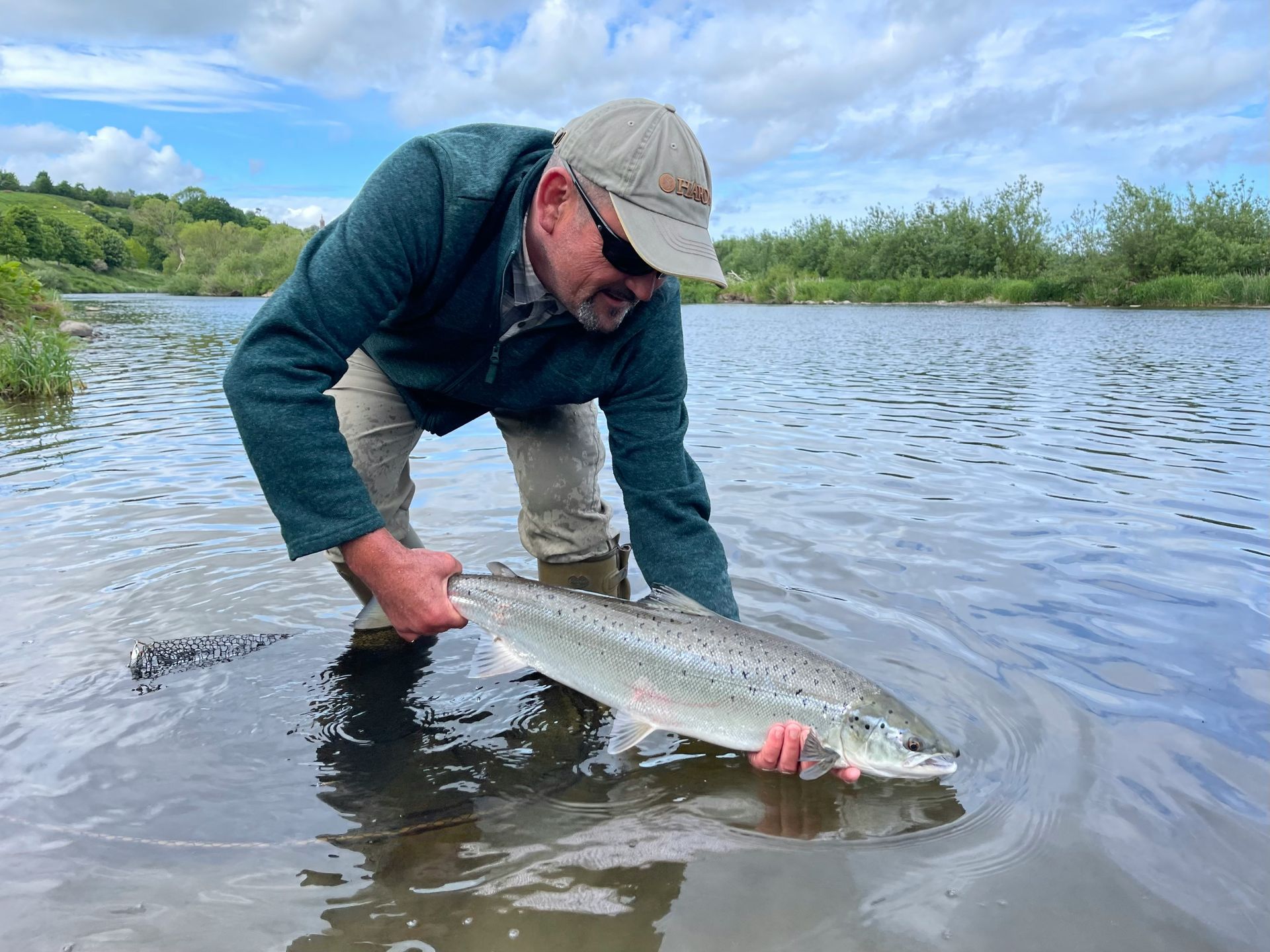 Man releasing a large silver fish into a calm lake under a cloudy sky