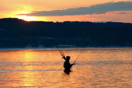 Silhouette of a person fishing in a calm lake at sunset, with orange reflections on the water.