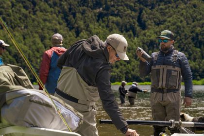 People in outdoor gear working beside a river, with mountains and trees in the background.