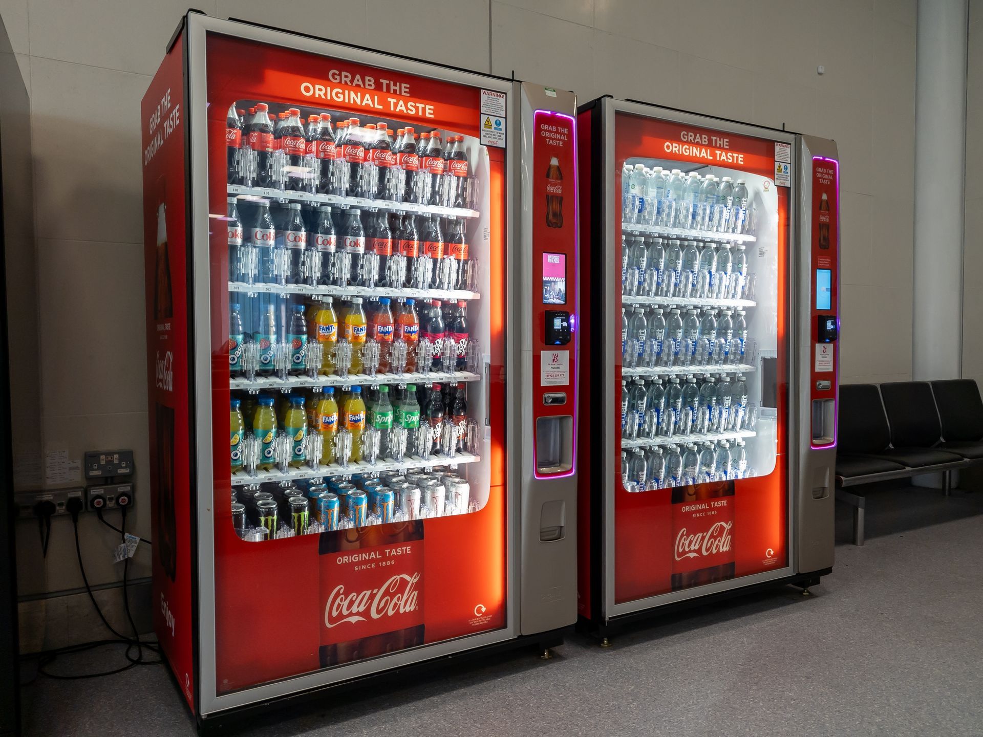 Two red Coca-Cola vending machines stocked with drinks in a hallway.