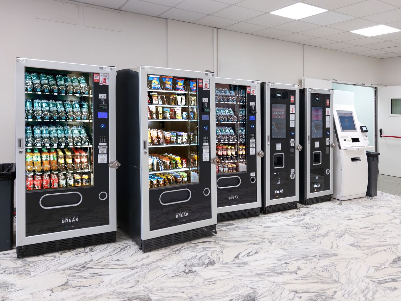 Row of vending machines in a bright indoor lobby with drinks, snacks, and a payment kiosk