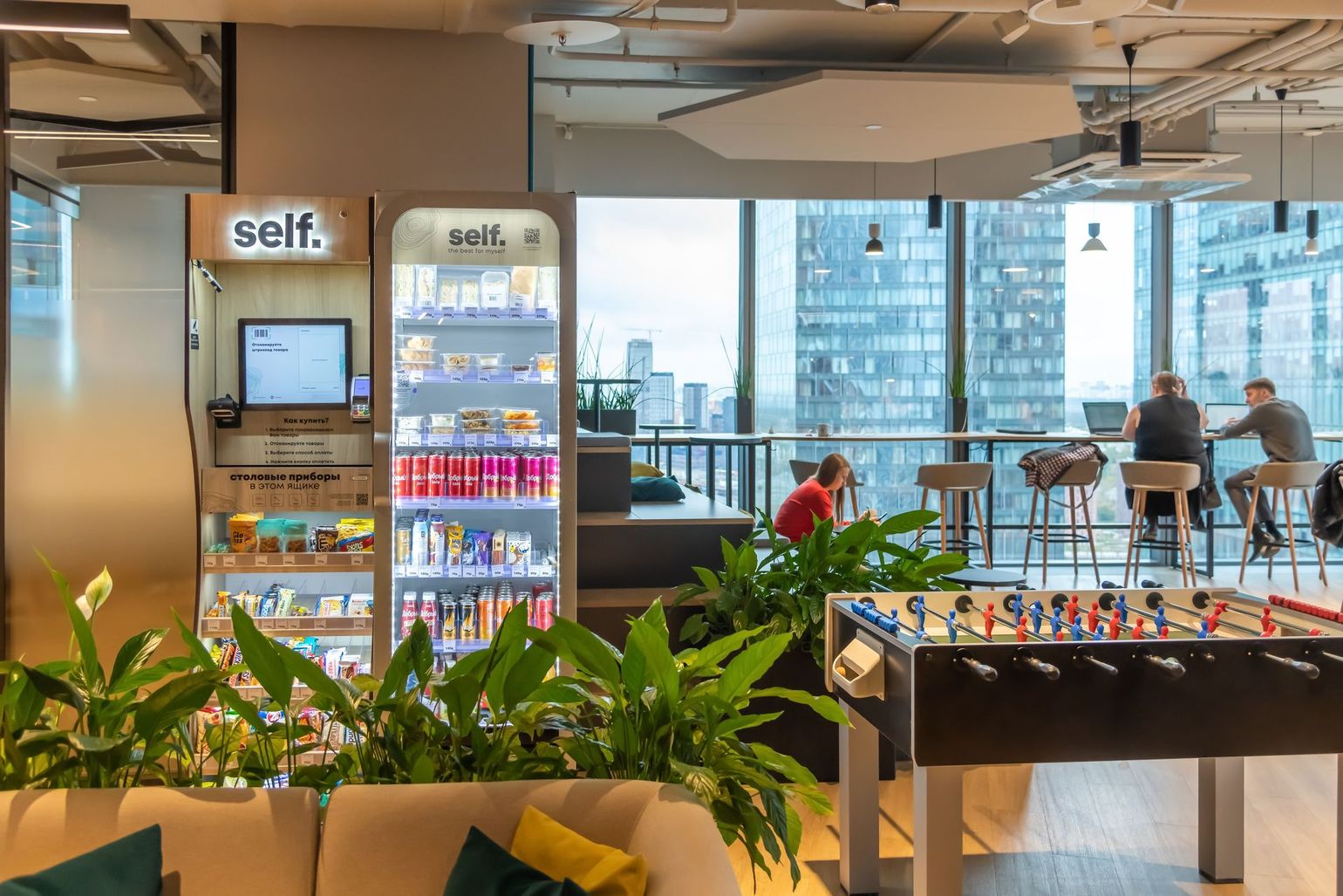 Modern lounge with plants, self-service fridges, and people seated by large windows overlooking the city