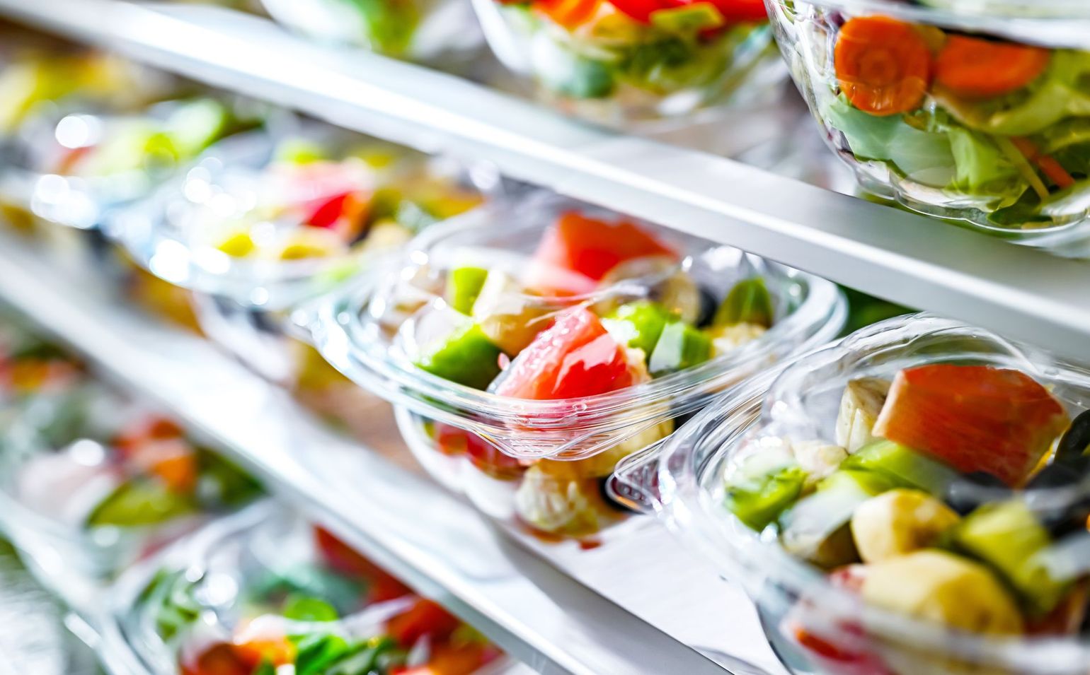 Clear plastic salad containers on a refrigerated grocery shelf, with colorful mixed greens and vegetables