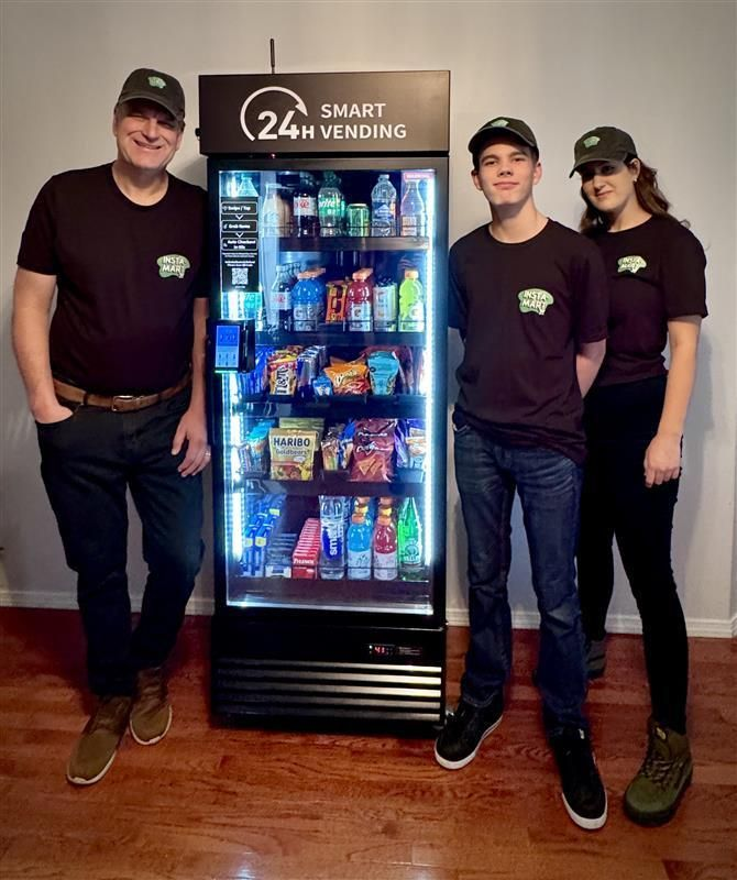 Three people stand beside a lit snack-and-drink vending machine in a hallway.