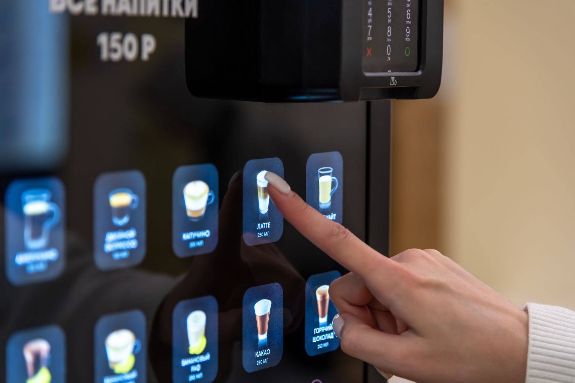 Person taps a touchscreen vending machine with glowing drink icons and a price label in Russian.