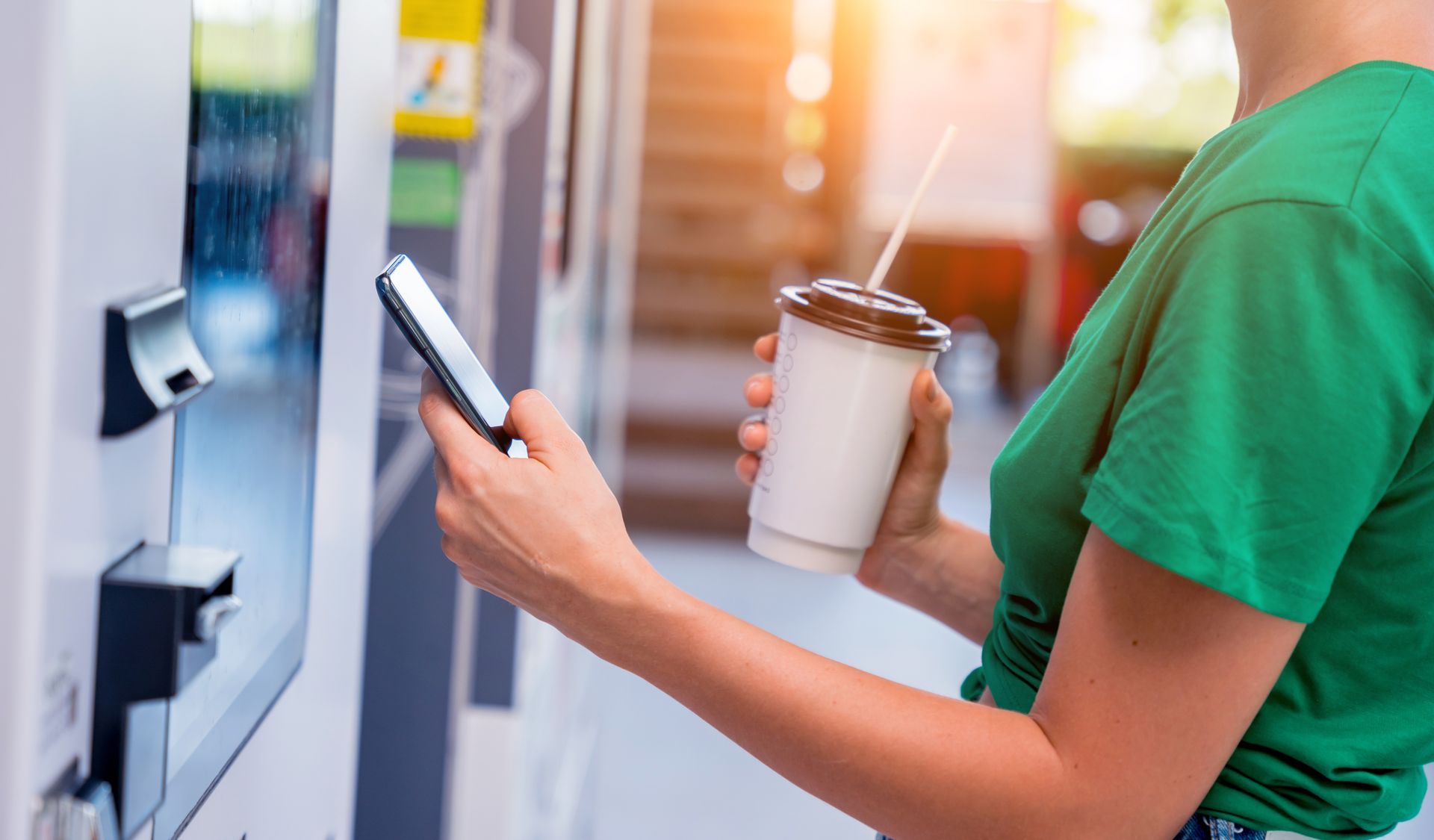 Person holding a smartphone and coffee cup at a vending machine, wearing a green shirt