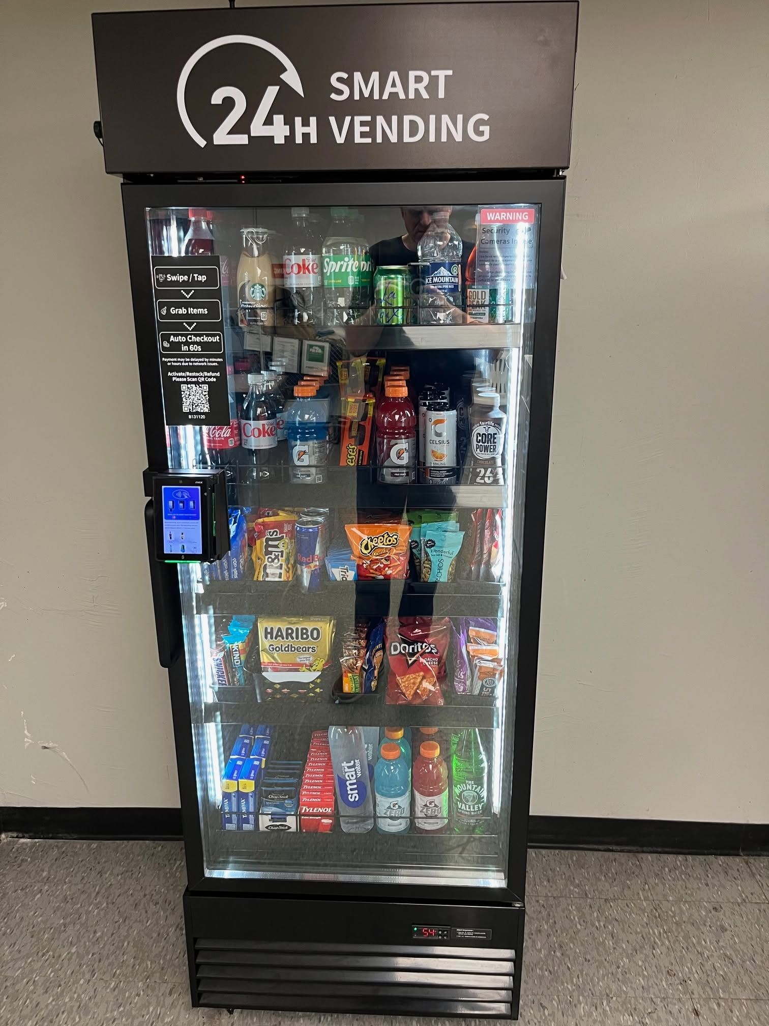 Smart vending machine with snacks and drinks, lit up indoors against a wall.