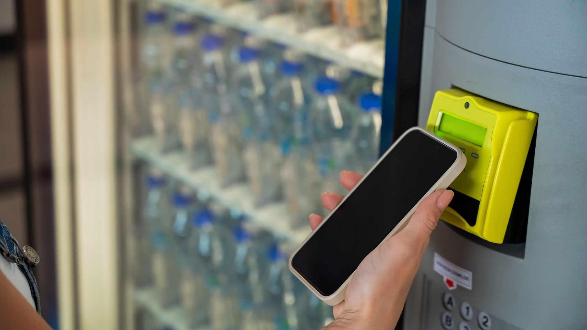 Hand holding a smartphone to a bright yellow vending machine payment scanner, with drink bottles in the background