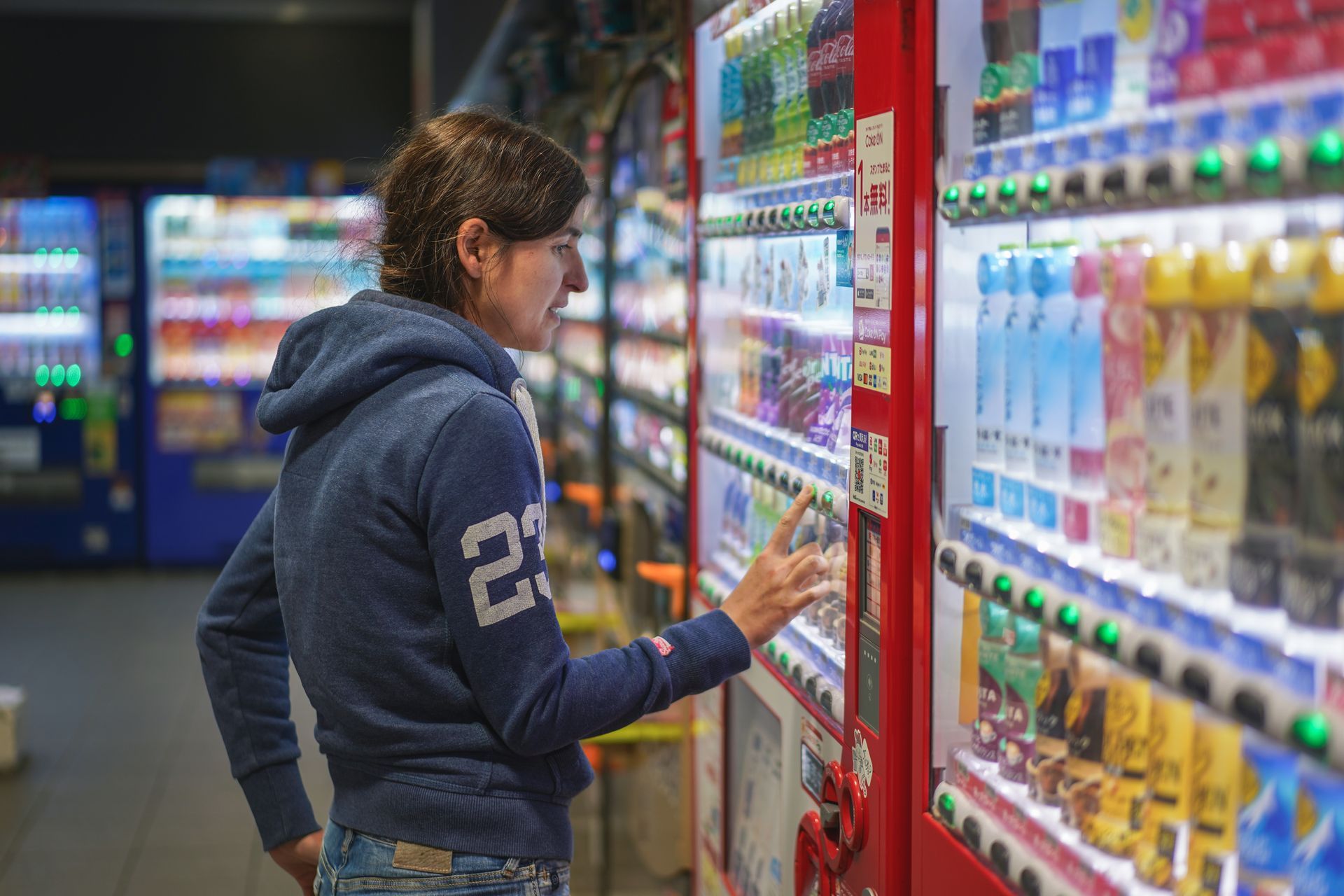 Person in a navy hoodie reaching for drinks in a store aisle with colorful vending machines.