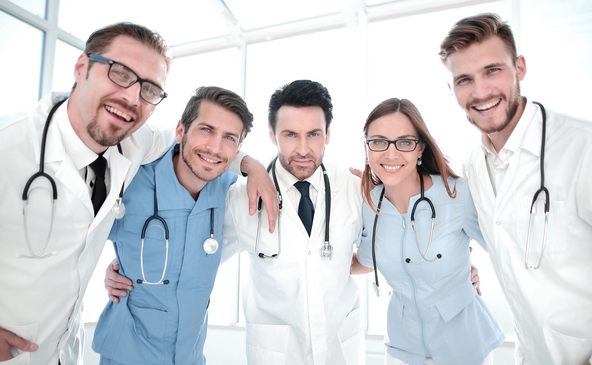 Cinque professionisti sanitari sorridenti, con indosso camici da laboratorio e stetoscopi, in un ambiente chiuso.