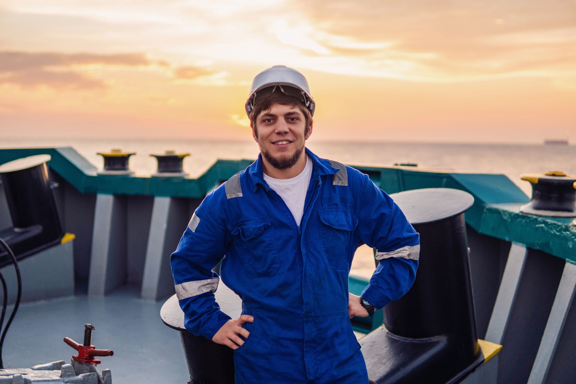 Persona sorridente con tuta blu e casco sul ponte di una nave al tramonto.