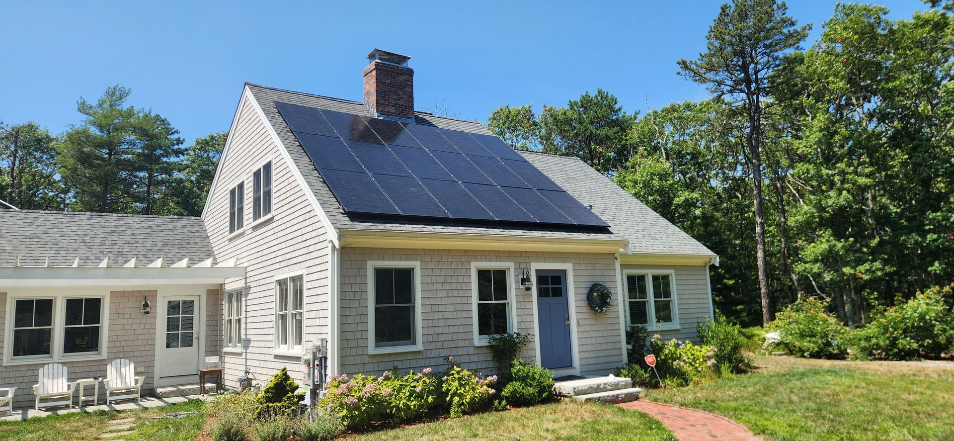 A light-colored house with solar panels on the roof, surrounded by trees and greenery under a clear blue sky.