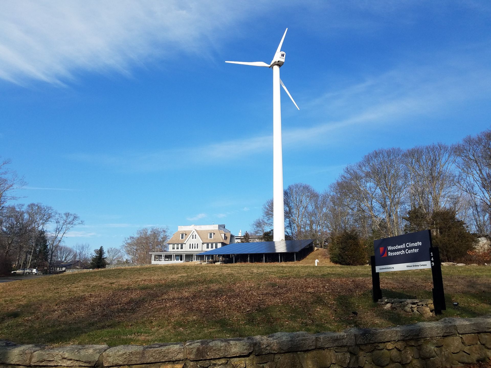 A large white wind turbine stands on a grassy hill next to a building, with solar panels at its base and a sign nearby.