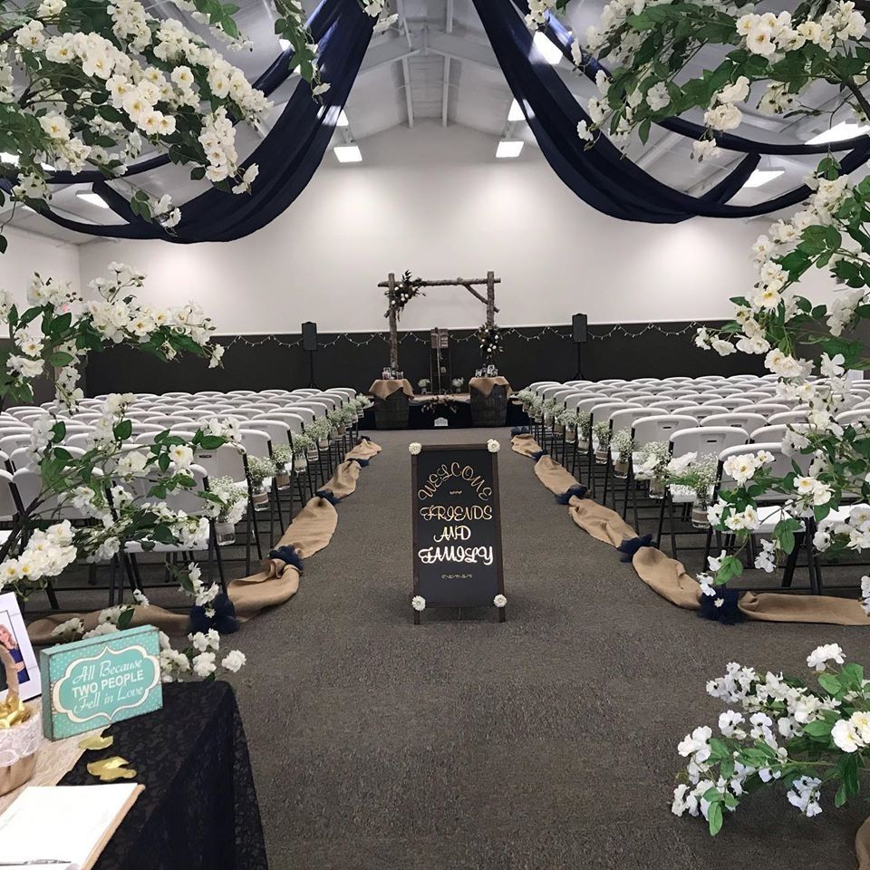 Flower-lined wedding aisle with white chairs and draped navy fabric under a tent.