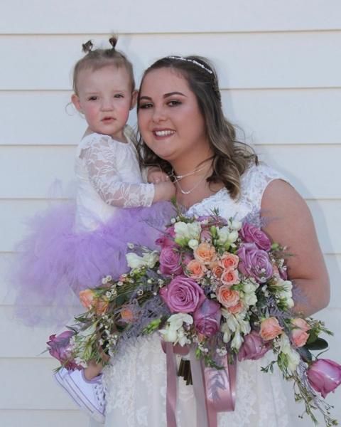 Woman holding a child beside a large pastel flower bouquet, both smiling against a white wall.