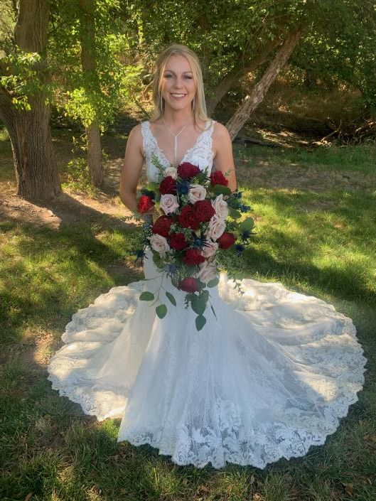 Bride in white lace gown holding a red and white bouquet outdoors under trees