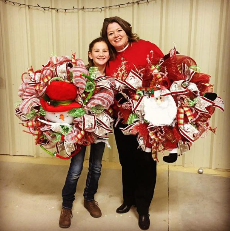 Two people holding large red-and-white decorative wreaths in front of beige curtains