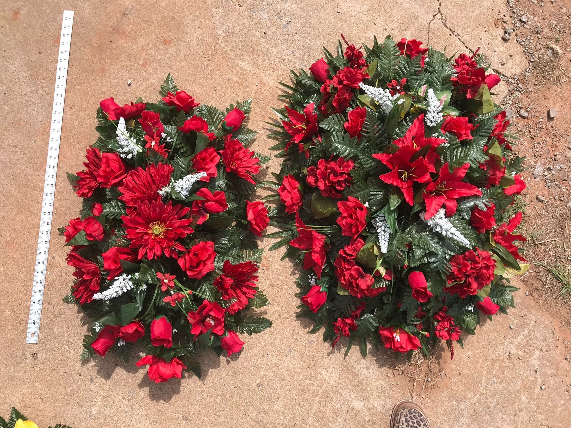 Two red-and-green floral wreaths on sandy ground, next to a measuring stick